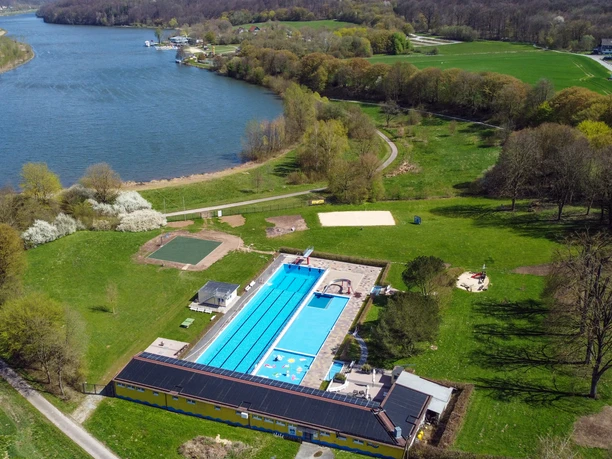 Das Luftbild zeigt ein Freibad mit einem blauen Schwimmbecken, umgeben von grünen Wiesen und Bäumen.