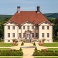 Vor einem bewaldeten Hügel erhebt sich ein elegantes Herrenhaus mit symmetrischer Gartenanlage.
