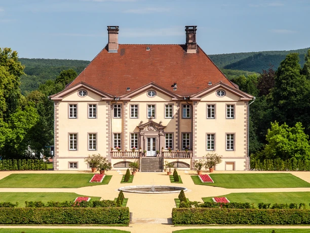 Schieder_Schloss_0024_RHa.jpg Vor einem bewaldeten Hügel erhebt sich ein elegantes Herrenhaus mit symmetrischer Gartenanlage.