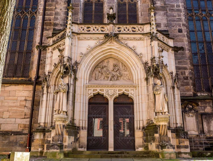 Tür der Stadtkirche St. Marien in Pirna Gotisches Kirchenportal mit zwei großen Holzflügeltüren, flankiert von steinernen Heiligenstatuen und einem Relief über dem Eingang.Gothic church portal with two large wooden double doors, flanked by stone statues of saints and a relief above the entrance.Gotický portál kostela se dvěma velkými dřevěnými dvoukřídlými dveřmi, po jejichž stranách jsou kamenné sochy světců a reliéf nad vchodem.Gotycki portal kościelny z dwoma dużymi drewnianymi podwójnymi drzwiami, otoczony kamiennymi posągami świętych i płaskorzeźbą nad wejściem.Gotisch kerkportaal met twee grote houten dubbele deuren, geflankeerd door stenen heiligenbeelden en een reliëf boven de ingang.Portale gotico della chiesa con due grandi porte doppie in legno, affiancate da statue di santi in pietra e da un rilievo sopra l'ingresso.