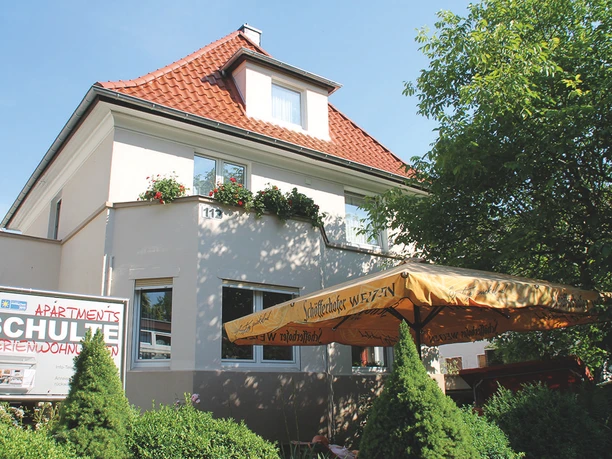 Ein helles, modernes Gebäude mit rotem Dach und Blumen an den Fenstern. Vor dem Haus befindet sich eine Terrasse mit einem Sonnenschirm.