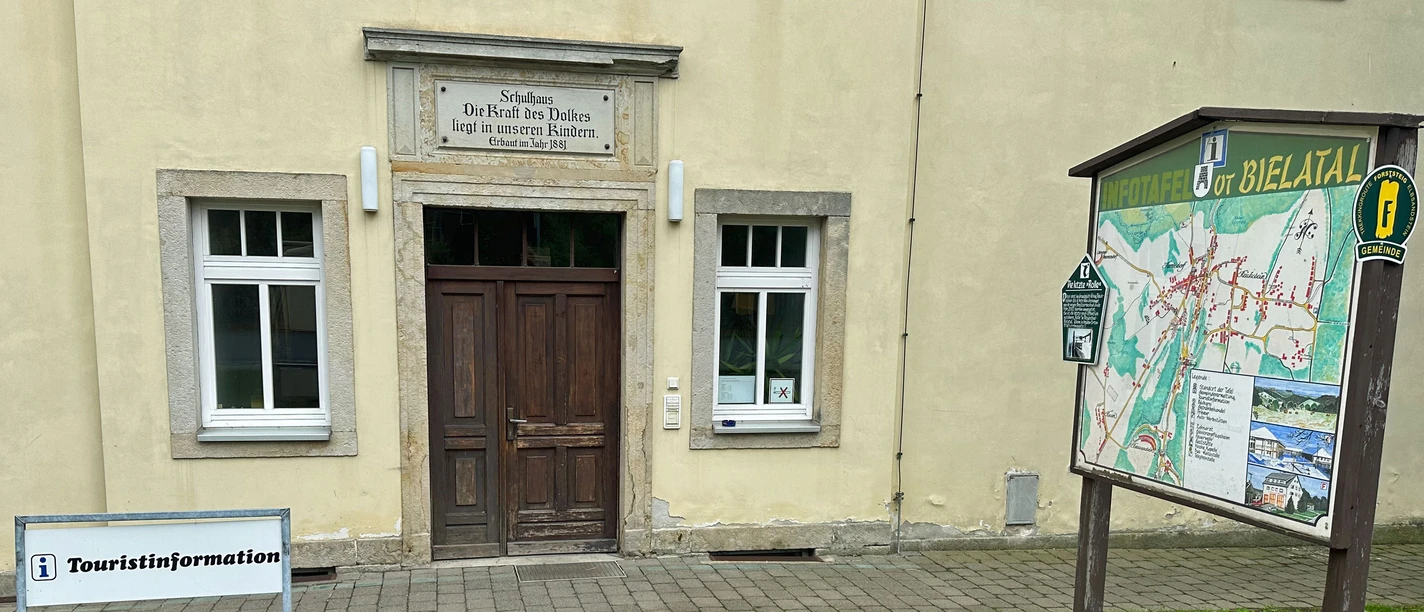 Ingang van een historisch gebouw met de inscriptie "De kracht van het volk ligt in onze kinderen", daarnaast een informatiebord en een bord "Toeristische informatie".
