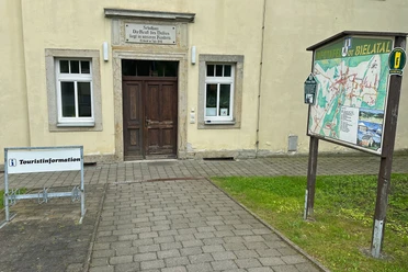 Eingang zur Touristinformation Rosenthal-Bielatal in der Gemeindeverwaltung Eingang eines historischen Gebäudes mit der Aufschrift "Die Kraft des Volkes liegt in unseren Kindern", daneben eine Infotafel und ein Schild "Touristinformation".