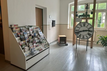 A room with a brochure rack full of brochures on the left and a large grandfather clock with Roman numerals on the right, with a plant next to it.