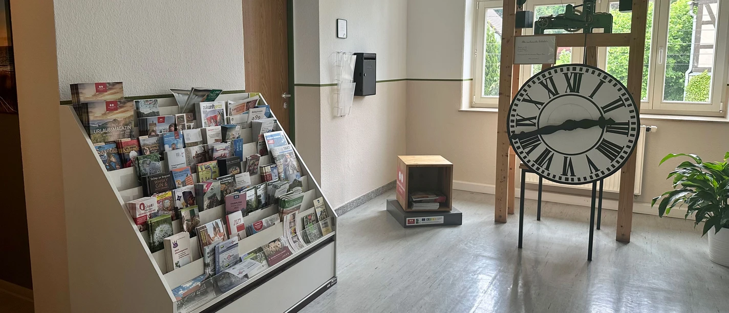 Een kamer met links een folderrek vol brochures en rechts een grote staande klok met Romeinse cijfers, met een plant ernaast.