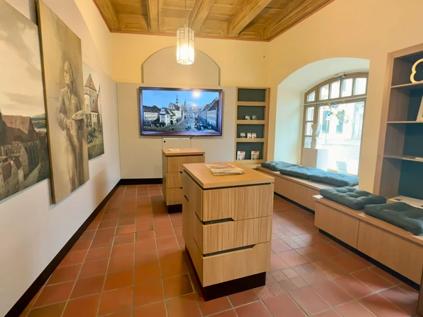 Bright room with wooden furniture, paintings on the wall, a large screen and a window seat with cushions; cozy, inviting atmosphere.
