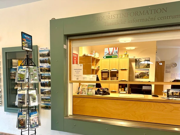 Ontmoetingsplaats Königstein Toeristische informatie met balie en brochurestand; lichtgekleurd houten meubilair, informatiemateriaal en een bord voor toiletgebruik zijn zichtbaar.