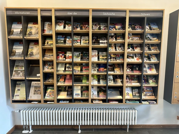 Prospektmaerial im Treffpunkt Königstein Ein Regal mit verschiedenen Broschüren und Flyern über die Sächsische Schweiz, sortiert in Fächern, darunter ein weißer Heizkörper.