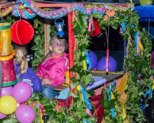 Fest der 1000 Laternen Das Bild zeigt zwei Kinder in einem bunt geschmückten Wagen, der mit grünen Blätterranken, bunten Bändern, Luftballons und Laternen dekoriert ist. Eines der Kinder trägt eine pinkfarbene Jacke und hat ein bunt geschminktes Gesicht. Das andere Kind trägt eine lila Jacke und ist von der Seite zu sehen. Die Szene wirkt fröhlich und erinnert an einen Festumzug oder ein Kinderfest.