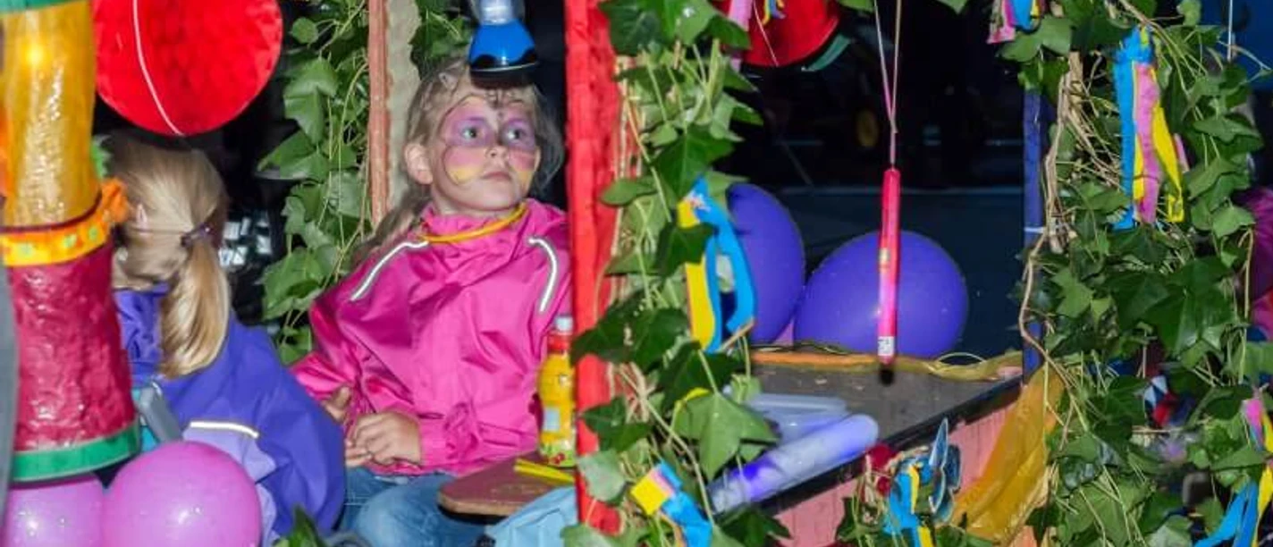 Fest der 1000 Laternen Das Bild zeigt zwei Kinder in einem bunt geschmückten Wagen, der mit grünen Blätterranken, bunten Bändern, Luftballons und Laternen dekoriert ist. Eines der Kinder trägt eine pinkfarbene Jacke und hat ein bunt geschminktes Gesicht. Das andere Kind trägt eine lila Jacke und ist von der Seite zu sehen. Die Szene wirkt fröhlich und erinnert an einen Festumzug oder ein Kinderfest.