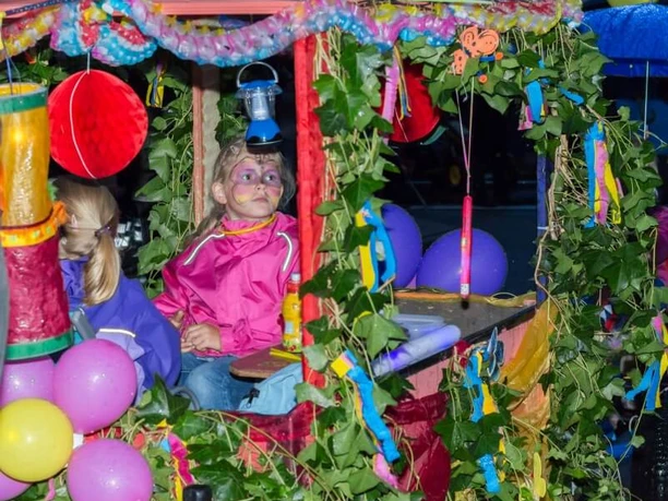 Fest der 1000 Laternen Das Bild zeigt zwei Kinder in einem bunt geschmückten Wagen, der mit grünen Blätterranken, bunten Bändern, Luftballons und Laternen dekoriert ist. Eines der Kinder trägt eine pinkfarbene Jacke und hat ein bunt geschminktes Gesicht. Das andere Kind trägt eine lila Jacke und ist von der Seite zu sehen. Die Szene wirkt fröhlich und erinnert an einen Festumzug oder ein Kinderfest.