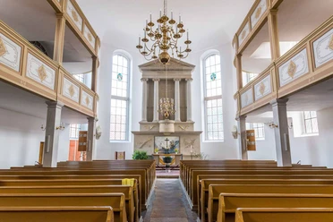 Interior view of a church with wooden benches, an altar with a cross and a large chandelier; bright, calm atmosphere.