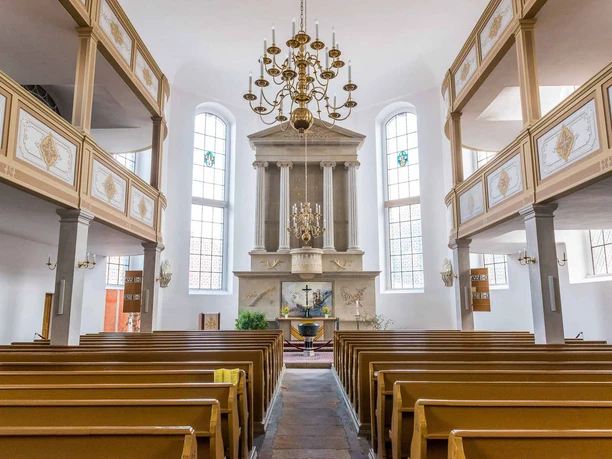 Binnenzicht van een kerk met houten banken, een altaar met een kruis en een grote kroonluchter; heldere, rustige sfeer.
