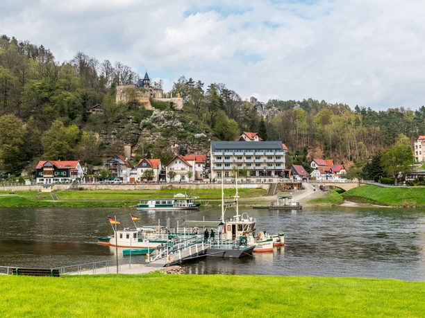Říční krajina s přívozem v popředí, za ním vesnice s barevnými domy a zříceninou hradu na zalesněném kopci.