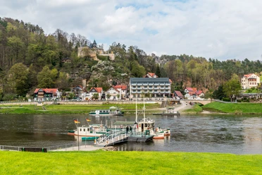 Fähre Kurort Rathen Flusslandschaft mit Fähre im Vordergrund, dahinter ein Dorf mit bunten Häusern und einer Burgruine auf einem bewaldeten Hügel.