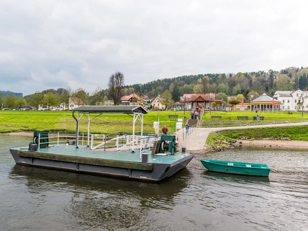 Zelený přívoz a malá loďka na řece, v pozadí zelená louka s chodci a domy pod zataženou oblohou.