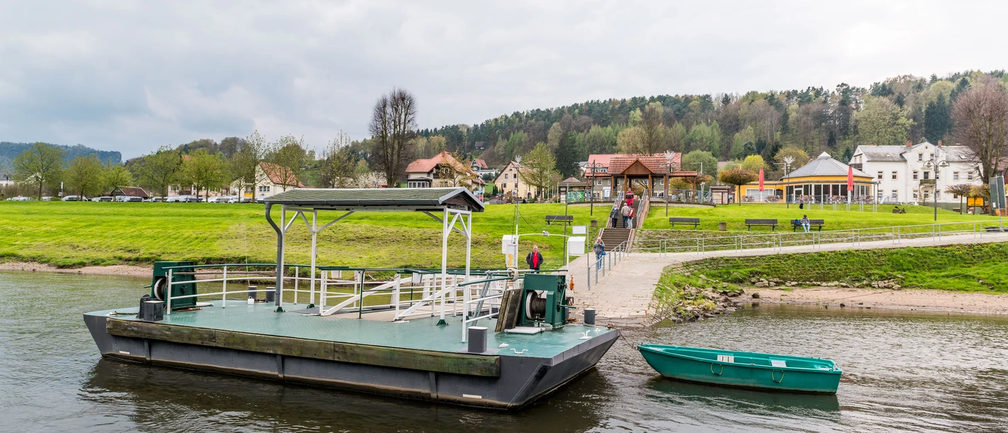 Eine grüne Fähre und ein kleines Boot auf einem Fluss, im Hintergrund eine grüne Wiese mit Spaziergängern und Häusern unter bewölktem Himmel.