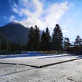 Eisplatz Farchant Eisplatz im Sonnenlicht, im Hintergrund Tannen und der Wank, mit aufsteigenden Wolken, rechts im Hintergrund eine Flutlichtlampe
