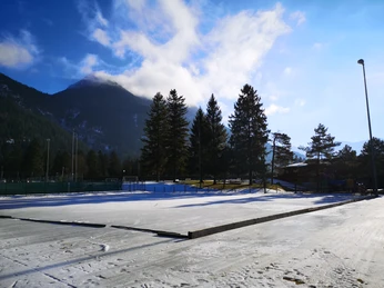 Eisplatz Farchant Eisplatz im Sonnenlicht, im Hintergrund Tannen und der Wank, mit aufsteigenden Wolken, rechts im Hintergrund eine Flutlichtlampe