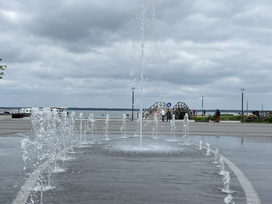 Springbrunnen und Strandterrassen am Seeufer, im Hintergrund bewölkter Himmel.