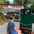 Feldbahn am Piesberg Kind fährt in grünem Feldbahnwagen durch grüne Landschaft am Piesberg mit Schiefergebäude hinten.