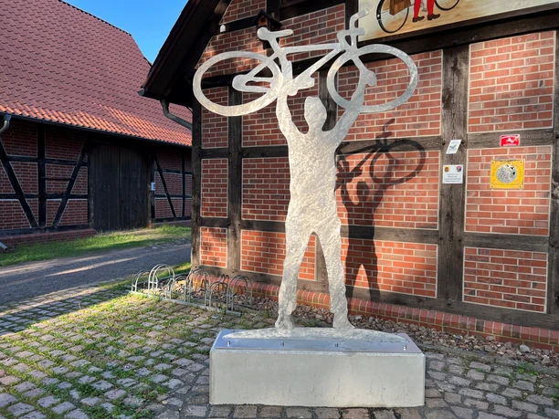 Sculpture of a person holding up a bicycle in front of a half-timbered building made of red brick.