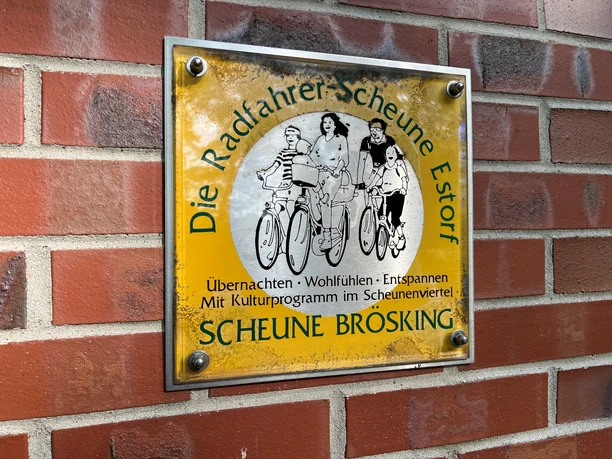 Metal sign on a brick wall with picture of family on bicycles and text: Radfahrerscheune Estorf.