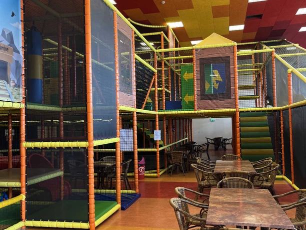 Western Joe.jpeg Großer Indoorspielplatz mit buntem Kletterturm, Rutschen und Sitzbereich mit Korbstühlen für Familien.