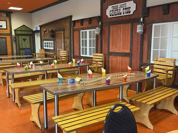 Western Joe.jpeg Bunte Sitzbänke und Tische im Westernstil mit Holzfassaden in einem kindgerechten Spielbereich.