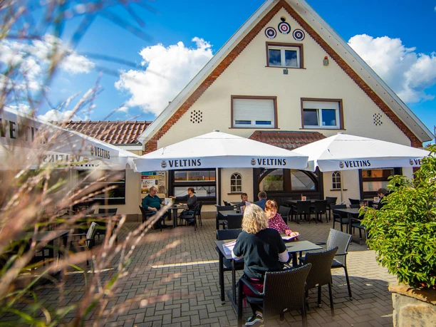 Biergarten.jpg Gäste sitzen unter weißen Sonnenschirmen im Außenbereich eines Restaurants bei sonnigem Wetter.