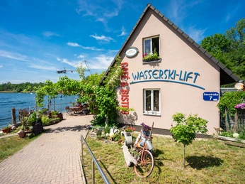 Wasserskilift, Gastonomie und Café am Kulkwitzer See Ein idyllisches Haus mit großer Terrasse am Kulkwitzer See, umgeben von blühenden Pflanzen. Der Wasserskilift im Hintergrund bietet sportliche Möglichkeiten in malerischer Kulisse. Ein Fahrrad mit Blumenschmuck verleiht dem Ambiente eine gemütliche Note.An idyllic house with a large terrace on Lake Kulkwitz, surrounded by flowering plants. The water ski lift in the background offers sporting opportunities in a picturesque setting. A bicycle decorated with flowers adds a cozy touch to the ambience.Idylický dům s velkou terasou u jezera Kulkwitz, obklopený kvetoucími rostlinami. Vlek na vodní lyže v pozadí nabízí sportovní vyžití v malebném prostředí. Útulnou atmosféru dotváří květinami vyzdobené jízdní kolo.Idylliczny dom z dużym tarasem nad jeziorem Kulkwitz, otoczony kwitnącymi roślinami. Wyciąg nart wodnych w tle oferuje możliwości uprawiania sportu w malowniczej scenerii. Rower ozdobiony kwiatami dodaje przytulnego akcentu do atmosfery.Een idyllisch huis met een groot terras aan het Kulkwitzmeer, omgeven door bloeiende planten. De waterskilift op de achtergrond biedt sportieve mogelijkheden in een schilderachtige omgeving. Een met bloemen versierde fiets zorgt voor een gezellige sfeer.Una casa idilliaca con un'ampia terrazza sul lago Kulkwitz, circondata da piante fiorite. L'impianto di risalita per lo sci nautico sullo sfondo offre opportunità sportive in un ambiente pittoresco. Una bicicletta decorata con fiori aggiunge un tocco accogliente all'ambiente.