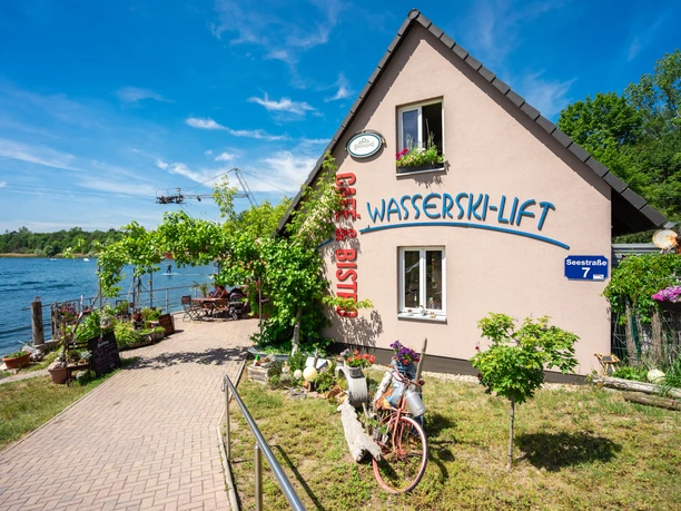 Water ski lift, restaurant and café on Lake Kulkwitz An idyllic house with a large terrace on Lake Kulkwitz, surrounded by flowering plants. The water ski lift in the background offers sporting opportunities in a picturesque setting. A bicycle decorated with flowers adds a cozy touch to the ambience.