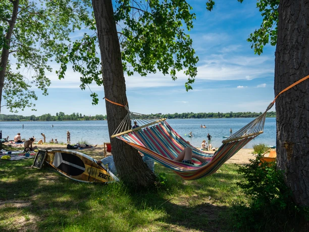 Entspannt abhängen am Kulkwitzer See A colorful hammock hangs between two trees on the shore of Lake Kulkwitz, while people swim in the water and relax on the beach.