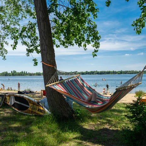 Kolorowy hamak wisi między dwoma drzewami na brzegu jeziora Kulkwitz, podczas gdy ludzie pływają w wodzie i relaksują się na plaży.