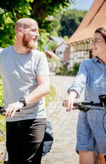 Bissendorf-Holte Zwei Personen schieben Fahrräder durch ein sonniges Dorf mit Fachwerkhäusern.