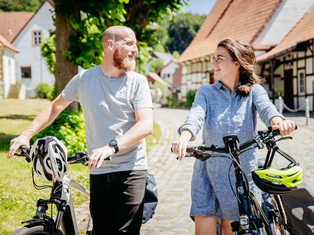 Zwei Personen schieben Fahrräder durch ein sonniges Dorf mit Fachwerkhäusern.