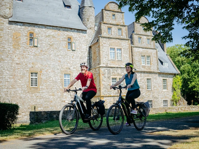 Die Schelenburg bei Bissendorf Zwei Radfahrer vor einem historischen steinernen Herrenhaus bei sonnigem Wetter.Two cyclists in front of a historic stone manor house in sunny weather.To cyklister foran en historisk herregård af sten i solskinsvejr.Twee fietsers voor een historisch stenen landhuis bij zonnig weer.