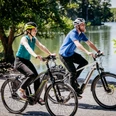 Rubbenbruchsee Zwei Radfahrer mit Schutzhelmen fahren fröhlich auf einem sonnigen Weg entlang eines Sees.Two cyclists wearing safety helmets ride happily along a sunny path beside a lake.To cyklister med sikkerhedshjelm kører glade ad en solrig sti langs en sø.Twee fietsers met veiligheidshelmen rijden vrolijk over een zonnig pad langs een meer.
