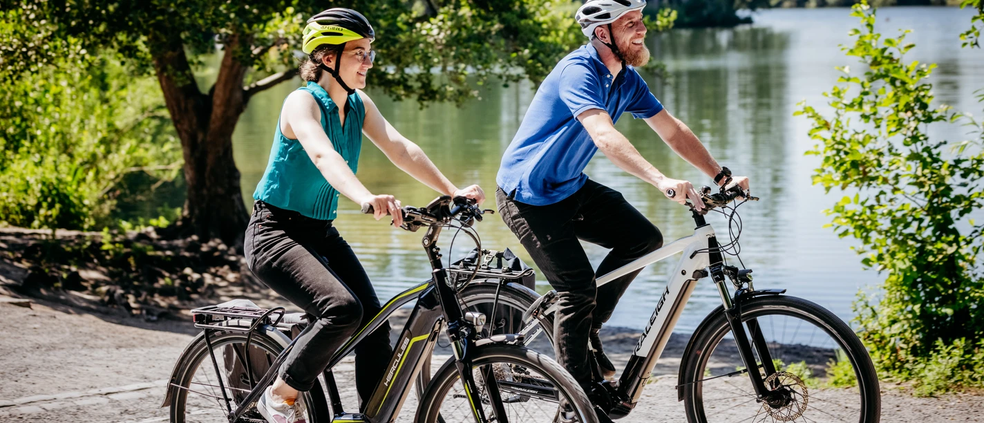 Twee fietsers met veiligheidshelmen rijden vrolijk over een zonnig pad langs een meer.