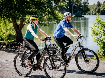 Rubbenbruchsee Zwei Radfahrer mit Schutzhelmen fahren fröhlich auf einem sonnigen Weg entlang eines Sees.Two cyclists wearing safety helmets ride happily along a sunny path beside a lake.To cyklister med sikkerhedshjelm kører glade ad en solrig sti langs en sø.Twee fietsers met veiligheidshelmen rijden vrolijk over een zonnig pad langs een meer.