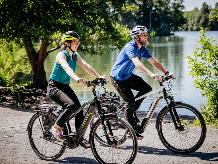 Rubbenbruchsee Zwei Radfahrer mit Schutzhelmen fahren fröhlich auf einem sonnigen Weg entlang eines Sees.Two cyclists wearing safety helmets ride happily along a sunny path beside a lake.To cyklister med sikkerhedshjelm kører glade ad en solrig sti langs en sø.Twee fietsers met veiligheidshelmen rijden vrolijk over een zonnig pad langs een meer.