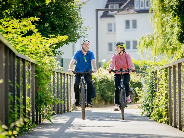 Der Haseuferweg in Osnabrück Twee fietsers met helmen rijden over een voetgangersbrug omgeven door weelderig groen.