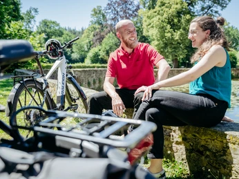 Die Schelenburg bei Bissendorf Lächelndes Paar sitzt auf einer Steinmauer, neben einem E-Bike im grünen Park.Smiling couple sitting on a stone wall, next to an e-bike in a green park.Et smilende par sidder på en stenmur ved siden af en elcykel i en grøn park.Lachend stel zittend op een stenen muur, naast een e-bike in een groen park.
