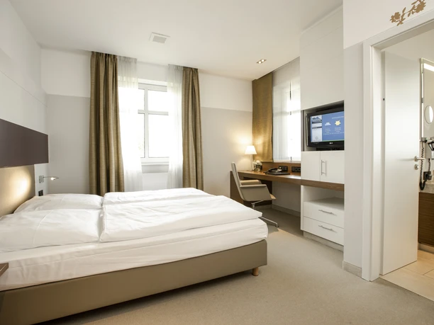 Doppelzimmer Hildegardhaus.jpg Modern eingerichtetes Hotelzimmer mit Doppelbett, Schreibtisch, Fernseher und angeschlossenem Bad.