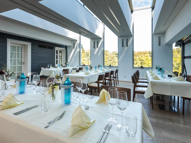 Pantholz 5.jpg Ein eleganter Speiseraum mit gedeckten Tischen, großen Fenstern und natürlichem Lichteinfall.
