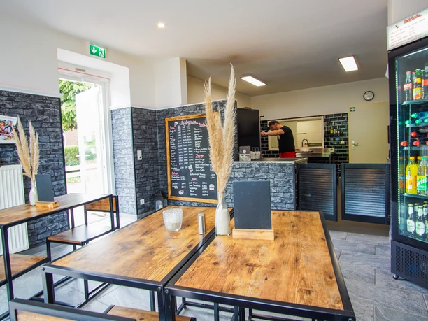 DSC07549-2.jpg Innenraum eines modernen Restaurants mit Holz-Metall-Tischen, schwarzer Steinoptik an den Wänden und einer gut sichtbaren Speisetafel. Im Hintergrund bereitet eine Person Speisen in der offenen Küche zu, rechts steht ein beleuchteter Kühlschrank mit Getränken.