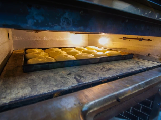 DSC07555-2.jpg Ein Backblech mit hell aufgegangenen Teiglingen befindet sich in einem beleuchteten Ofen. Die Oberfläche der Teigstücke glänzt durch die Hitze, während das Blech auf einem metallenen Rost liegt. Die Umgebung wirkt sauber und funktional, typisch für eine professionelle Küche.