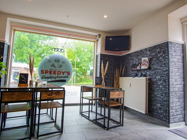 DSC07561-2 Heller Innenraum des Restaurants Speedy’s Amelinghausen mit großen Fenstern, modernem Mobiliar aus Holz und Metall sowie dunklen Steinwänden. Auf dem Fenster ist das Restaurantlogo zu sehen, draußen grünes Laub. Ein Fernseher hängt an der Wand, Dekoelemente sorgen für ein modernes Ambiente.