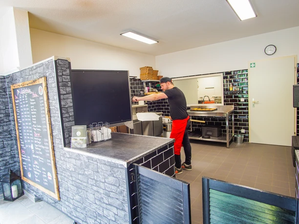 Ein Mitarbeiter formt Teig in einer modernen Pizzaküche Ein Mitarbeiter formt Teig in einer modernen Pizzaküche mit dunklen Fliesen und heller Beleuchtung. Im Vordergrund steht eine Theke mit Speisekarte, im Hintergrund liegen Pizzaschachteln und Küchenutensilien bereit. Die Umgebung wirkt sauber und barrierefrei gestaltet.