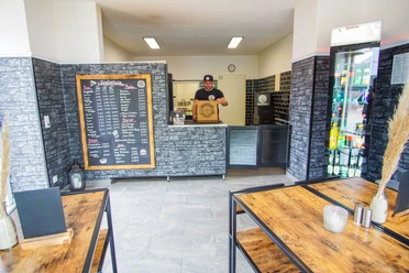 Innenraum von Speedy's Heller Gastraum mit modernen Holztischen, einer Getränkekühlvitrine und einer großen Menütafel an der Wand. Hinter der Theke steht ein Mitarbeiter mit einer Pizzaschachtel. Die Einrichtung wirkt klar strukturiert, barrierefrei zugänglich und auf Selbstbedienung ausgelegt.